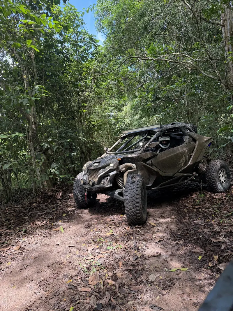 Mud-covered Can-Am Maverick R on a jungle trail, with a helmeted driver at the wheel