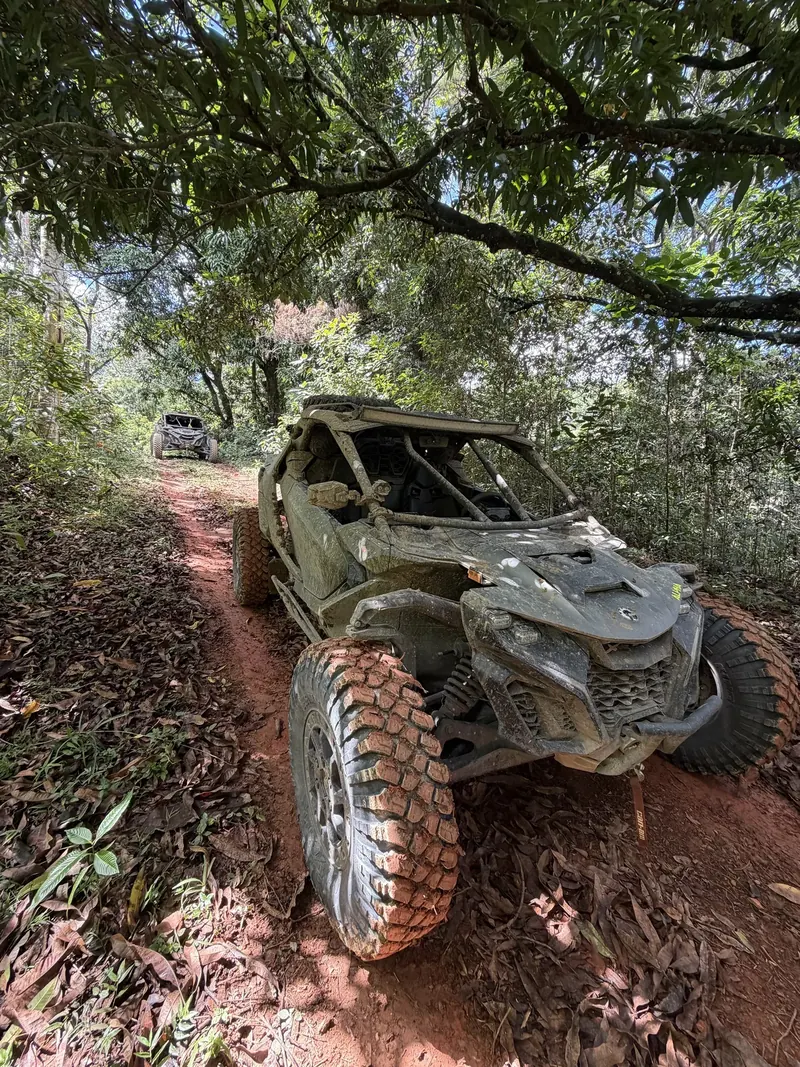 Mud-covered Can-Am Maverick R on a red dirt trail in the forest, with a second Can-Am behind