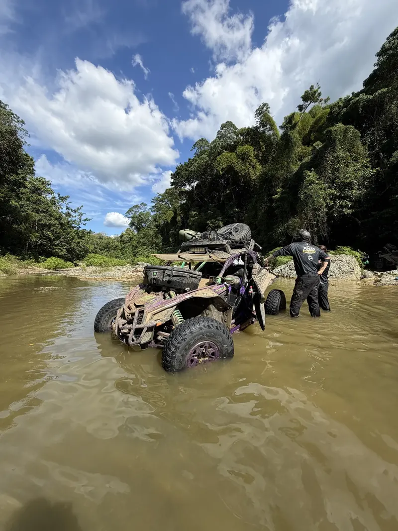 Purple Can-Am stopped in a deep river below jungle-covered hills, with two people standing in the water beside it